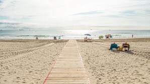 Ein hölzerner Steg führt zu einem Sandstrand mit Sonnenschirmen und Liegestühlen.