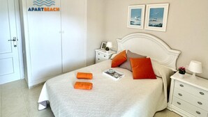Ein ordentlich bezogenes Bett mit weißer, strukturierter Bettdecke, zwei orangefarbenen Handtüchern und einem Tablett mit Tassen und einem Buch. An der Wand hängen zwei gerahmte Bilder, ein weißer Nachttisch mit Lampe und einer kleinen Pflanze, und ein weißer Kleiderschrank mit einem Schild darüber.