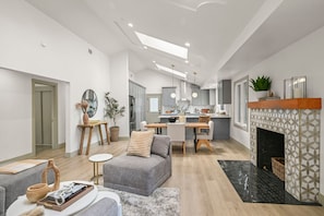 A modern living room with a grey sofa, a dining area with a wooden table, a kitchen with grey cabinets, and a fireplace with patterned tiles.