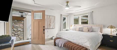 Un dormitorio con cama, mesitas de noche, un banco, ventilador de techo y puerta que da a un balcón.