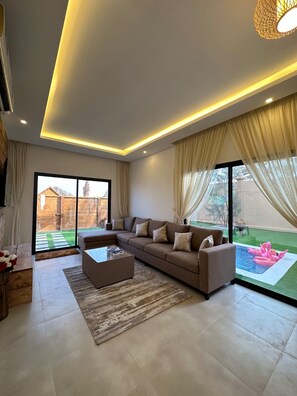 A modern living room with a sectional sofa, a coffee table, and a view of a backyard with a swimming pool. - Al Rayan Garden (Nabih saleh)