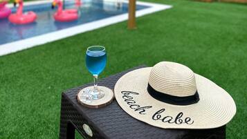 A backyard with a swimming pool, two pink flamingos, a drink, and a straw hat with the word "beach babe" on a table.