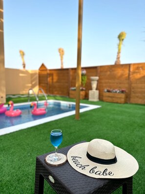 A backyard with a swimming pool, two pink flamingos, a drink, and a straw hat with the word "beach babe" on a table. - Al Rayan Garden (Nabih saleh)