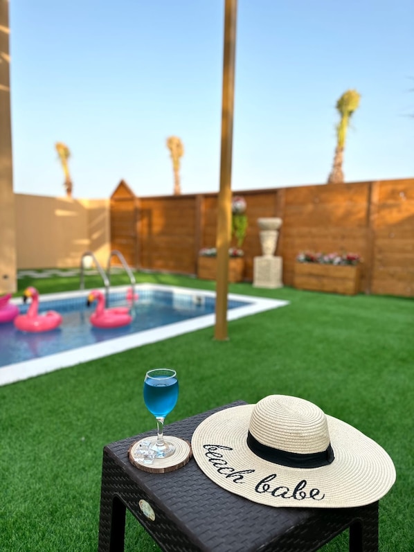 A backyard with a swimming pool, two pink flamingos, a drink, and a straw hat with the word "beach babe" on a table.