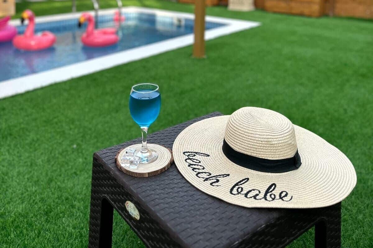 A backyard with a swimming pool, two pink flamingos, a drink, and a straw hat with the word "beach babe" on a table.