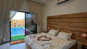 A bedroom with a wooden headboard, a bed with white linens, a heart-shaped decoration on the bed, and a view of a swimming pool through the glass door.