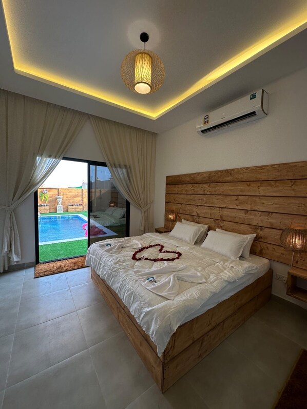A bedroom with a wooden headboard, a bed with white linens, a heart-shaped decoration on the bed, and a view of a swimming pool through the glass door. - Al Rayan Garden (Nabih saleh)