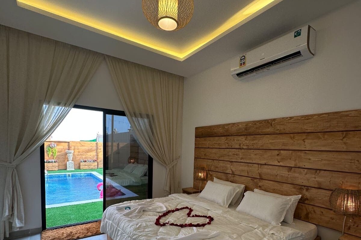 A bedroom with a wooden headboard, a bed with white linens, a heart-shaped decoration on the bed, and a view of a swimming pool through the glass door.