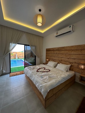 A bedroom with a wooden headboard, a bed with white linens, a heart-shaped decoration on the bed, and a view of a swimming pool through the glass door.