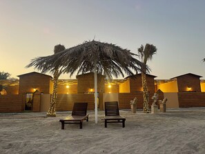 A beachside resort with wooden cabins, two lounge chairs under a palm tree, and decorative lights. - Al Rayan Garden (Nabih saleh)