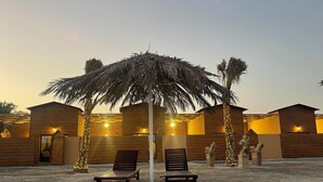 Resort à beira-mar com cabanas de madeira, duas espreguiçadeiras sob uma palmeira e iluminação decorativa.