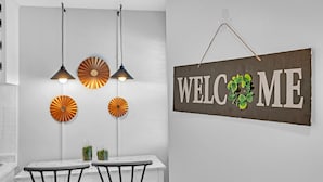 Un comedor con una mesa blanca y dos sillas, luces colgantes en el techo y un letrero de 'WELCOME' decorado con un arreglo floral.