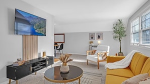 Una sala de estar moderna con un televisor de pantalla plana, un mueble de entretenimiento negro, un sofá amarillo y una zona de comedor con una silla y una mesa blancos.