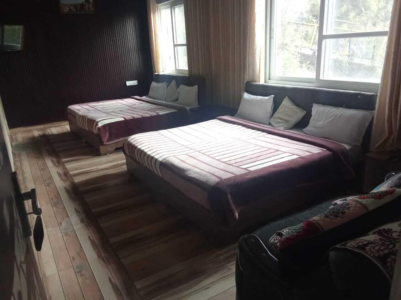 A room with two beds, wooden flooring, a window with curtains, and a view of greenery outside.