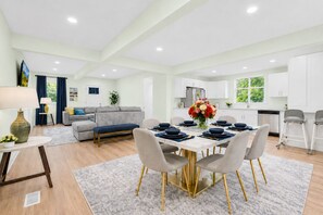 A modern kitchen with white cabinets, a dining area with a table set for four, and a living room with a grey sofa and a TV. - LAKE GEORGE VILLAGE. 1 Block to BEACH, SHEPHERDS PARK & CANADA St. PET  Friendly (Lake George)