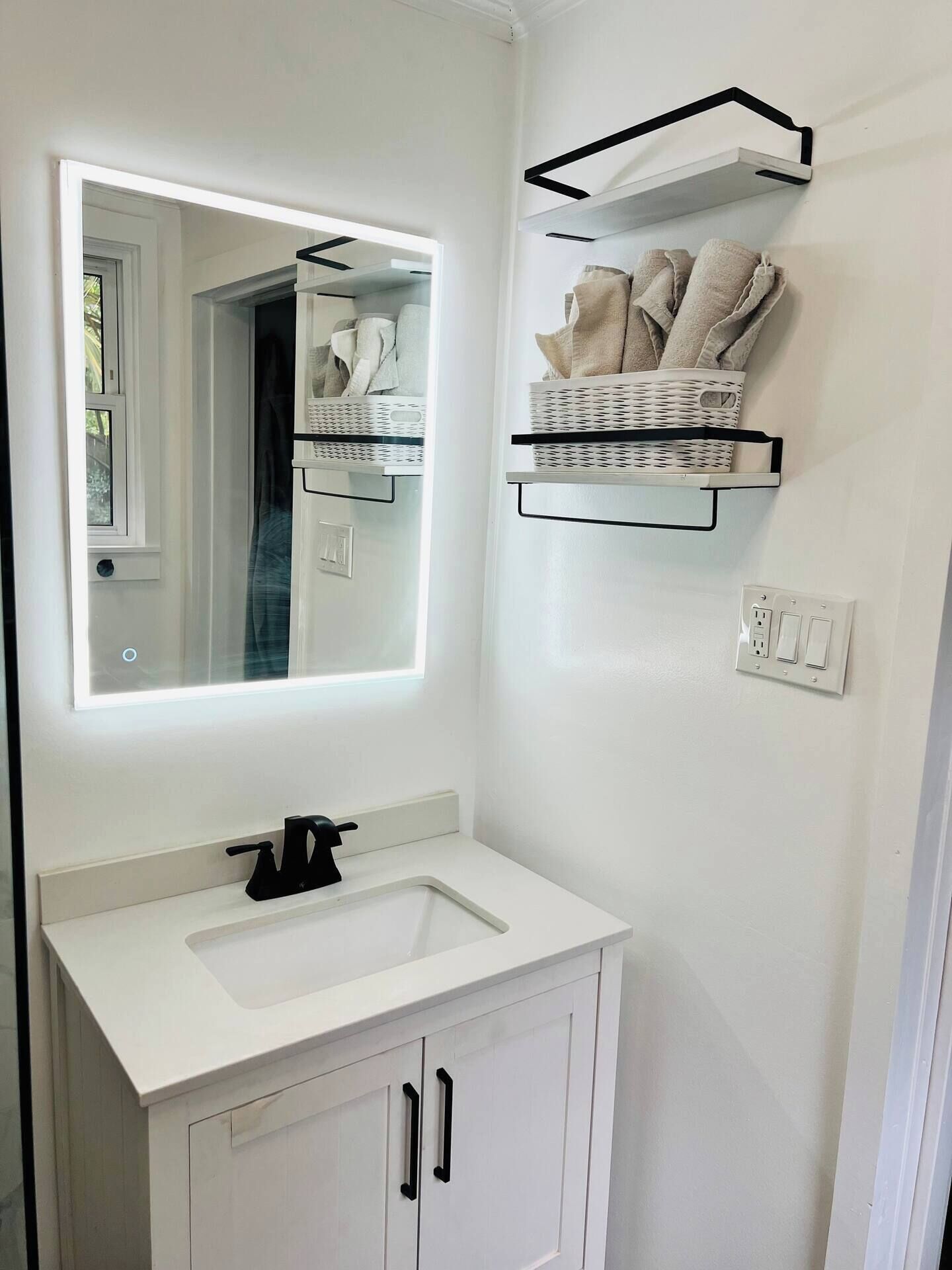 A bathroom with a white vanity, a mirror, a black faucet, and two white cabinets. There are two white wicker baskets on floating shelves above the vanity.