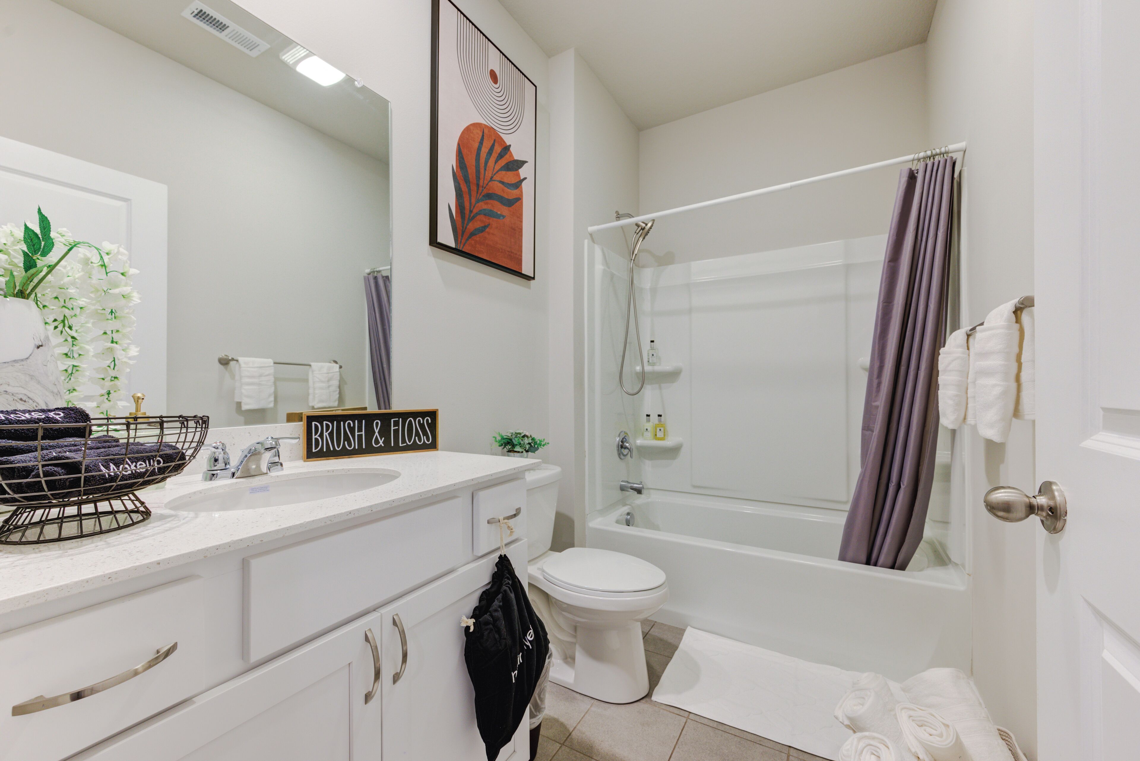 A modern bathroom with a white sink, toilet, and bathtub. There is a towel rack, a shower curtain, and a framed artwork on the wall.