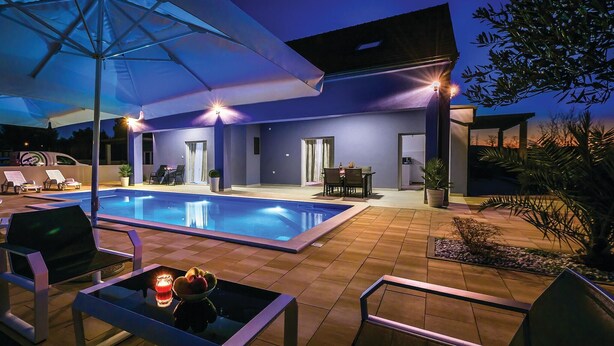 A modern outdoor pool area with lounge chairs and a dining table, surrounded by a patio with potted plants. - 6 bedroom amazing home in Bilice (Bilice)