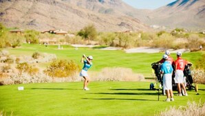 A golf course with a mountainous backdrop, a golfer in mid-swing, and a group of people standing nearby. - Large 3 Bedroom in Central Scottsdale w/ Pool, Clubhouse, Fitness Room and More… (Scottsdale)