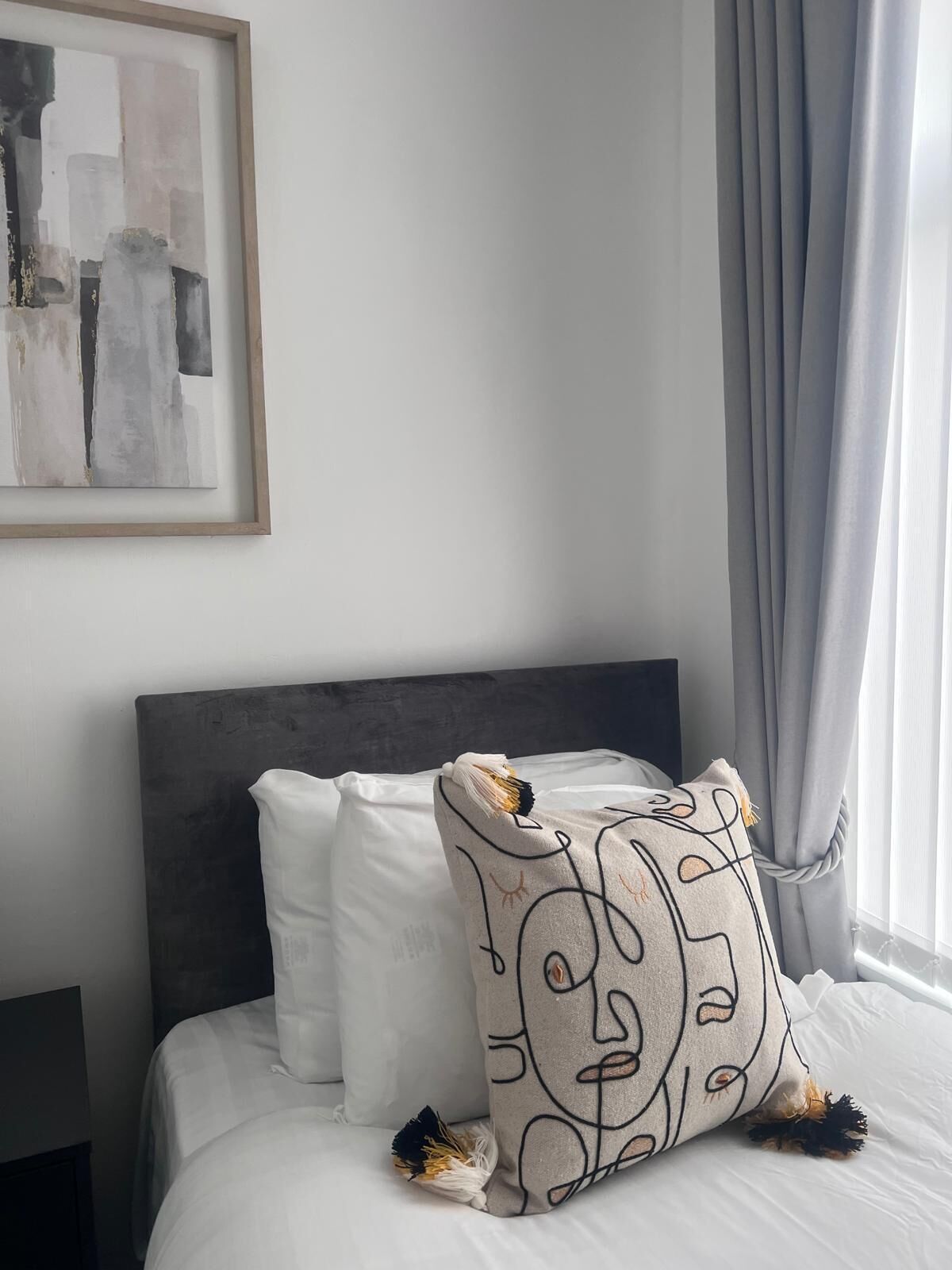 Un lit bien fait, avec une literie blanche et une tête de lit grise. Un coussin décoratif orné d’un motif représentant un visage est posé sur le lit.
