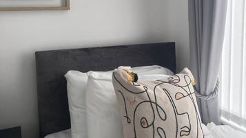 Un lit bien fait, avec une literie blanche et une tête de lit grise. Un coussin décoratif orné d’un motif représentant un visage est posé sur le lit.