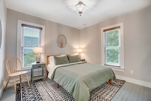 A bedroom with a bed, two bedside tables with lamps, a chair, a patterned rug, and a ceiling light fixture.