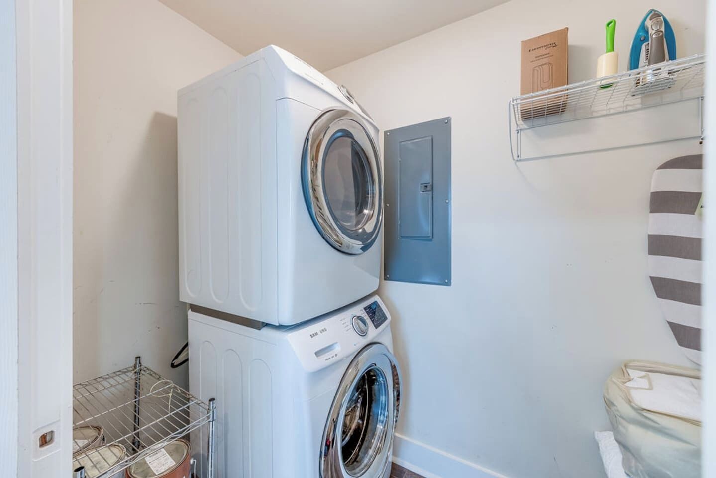 A compact laundry room with stacked washers and dryers, a metal drying rack, and a shelf with cleaning supplies.