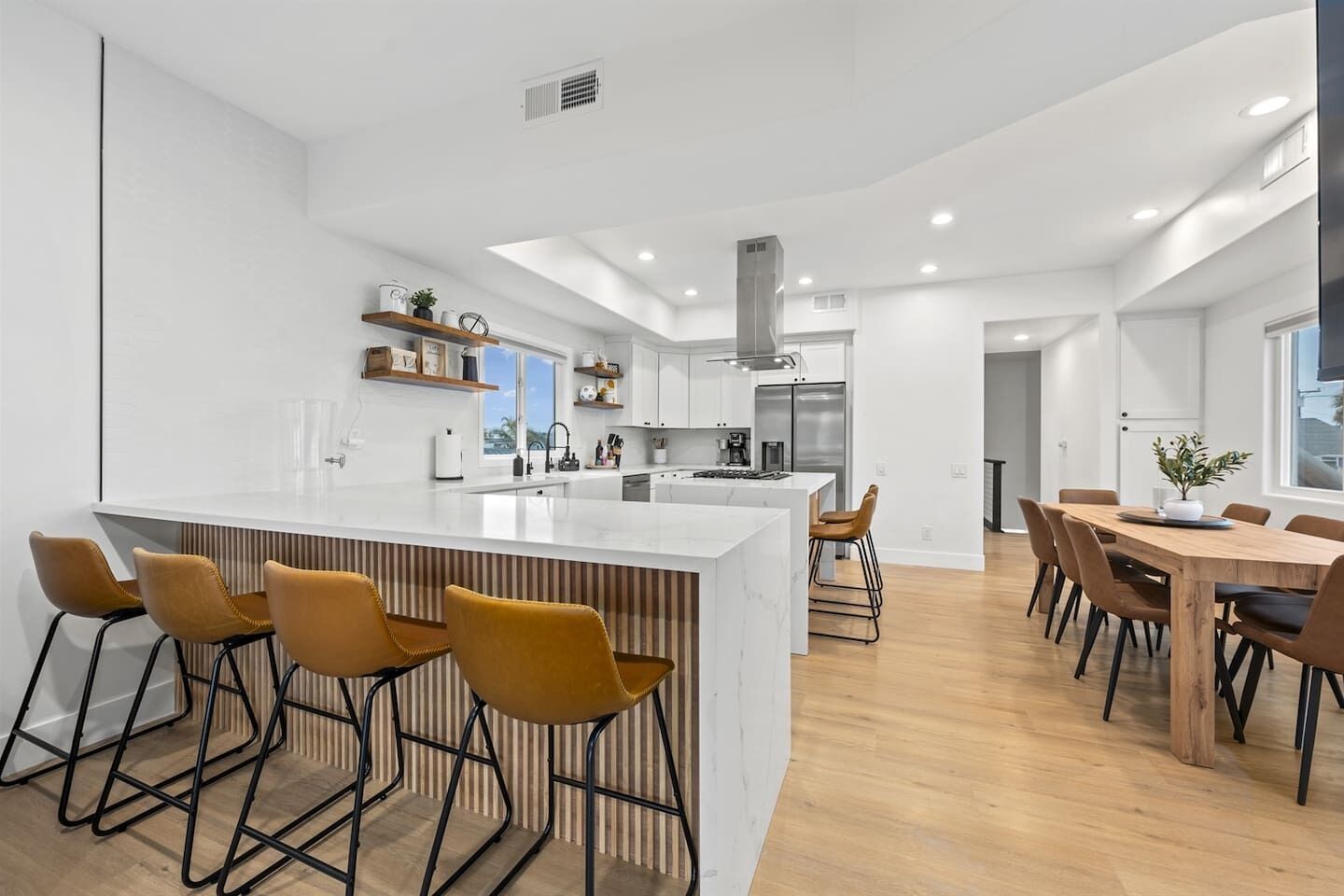 Una cocina moderna con una isla central, taburetes de bar y un comedor con mesas y sillas de madera.