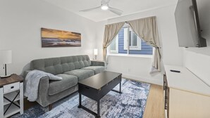 Ein modernes Wohnzimmer mit einem grauen Sofa, einem kleinen Tisch, einem Deckenventilator, einem Fernseher und einem Fenster mit Vorhängen.