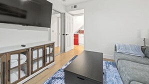 Ein modernes Wohnzimmer mit einem Flachbildfernseher, einer Holzkabine zur Aufbewahrung, einem grauen Sofa und einem blauen Teppich mit Muster.
