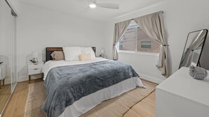 Ein Schlafzimmer mit Bett, Nachttischen, Deckenventilator, Fenster mit Vorhängen und einem Spiegel.