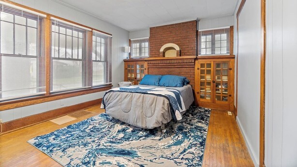 A bedroom with a bed, a brick fireplace, and large windows with blinds.