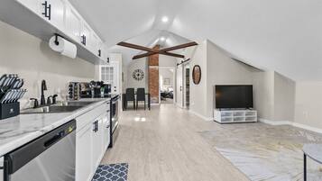 A modern kitchen with a dishwasher, a dining area with a brick accent wall, and a living room with a TV.