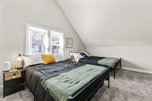 A bedroom with two beds, a window with blinds, a bedside table with a lamp, and a framed picture on the wall.