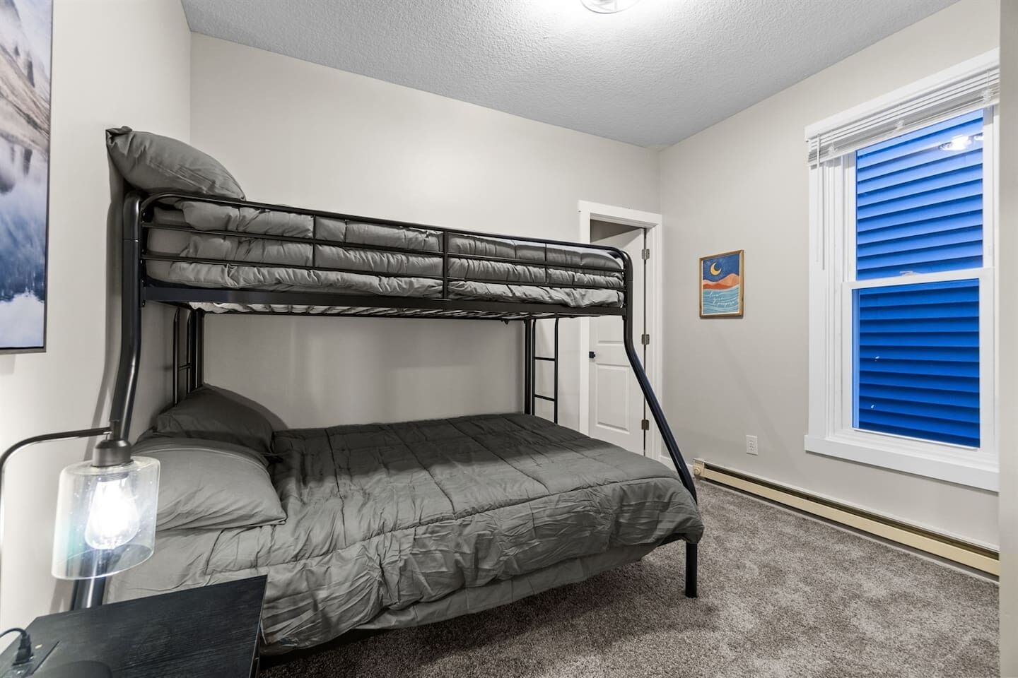 A bunk bed in a room with a desk, lamp, and a window with blue blinds.