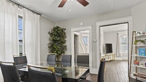 A modern living room with a glass-top dining table, black chairs, a ceiling fan, a large green plant, and a bookshelf with books and games. - Family Friendly Home for 8 (Fort Wayne)