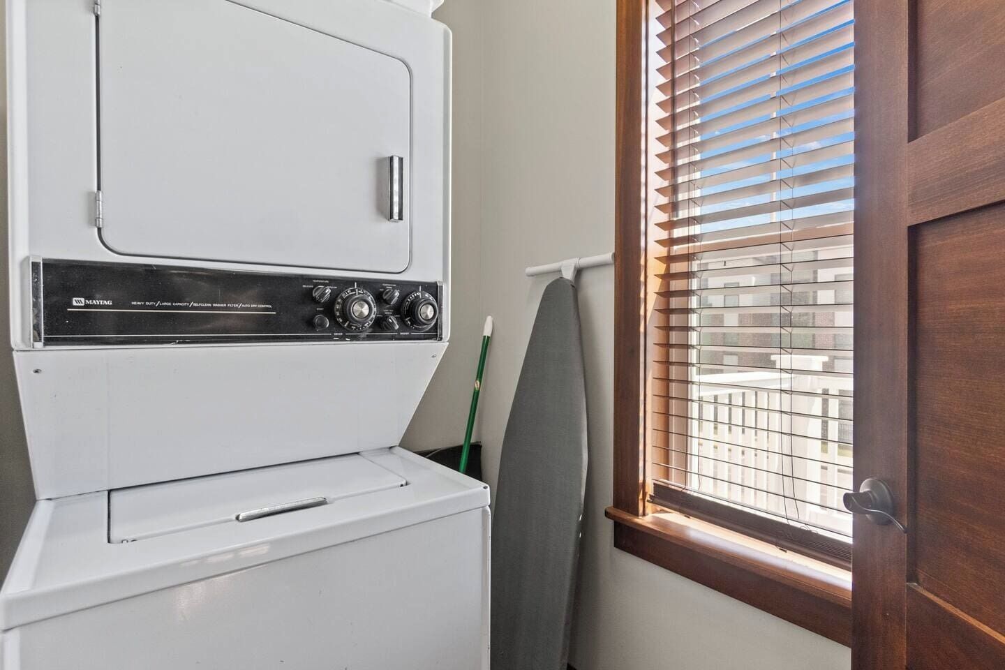A compact laundry area with a Maytag washer and dryer, a window with blinds, and a door.