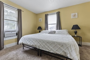 A bedroom with a bed, two bedside tables, a window with curtains, and a wall-mounted air conditioner.