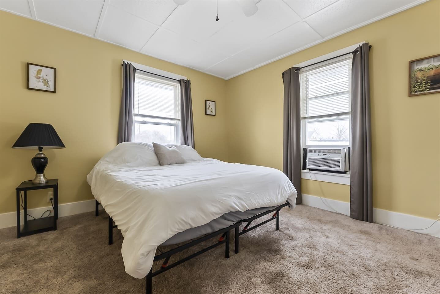 A bedroom with a bed, two windows with curtains, a bedside lamp, and a carpeted floor.