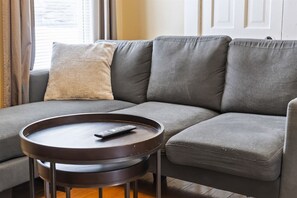 A gray sofa with a remote control on a round wooden table, a beige pillow, and a window with curtains.