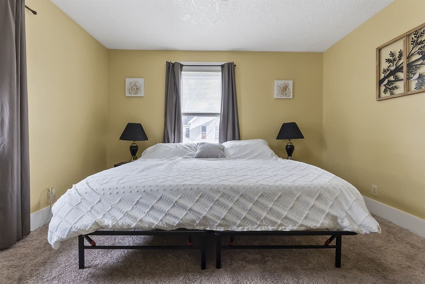A bedroom with a bed, two bedside lamps, a window with curtains, and two wall art pieces.