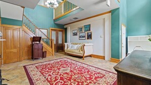 A spacious living area with a staircase, a large red patterned rug, a white sofa, and a chandelier.