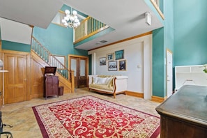 A spacious living area with a staircase, a large red patterned rug, a white sofa, and a chandelier.
