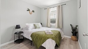 A bedroom with a bed, a bedside table with a lamp, a window with curtains, a wall-mounted decoration, and a potted plant.