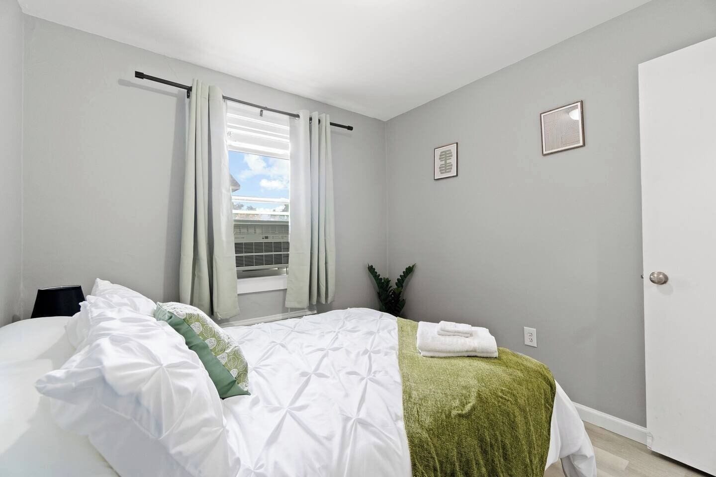 A bedroom with a bed, a window with curtains, a potted plant, and a door.