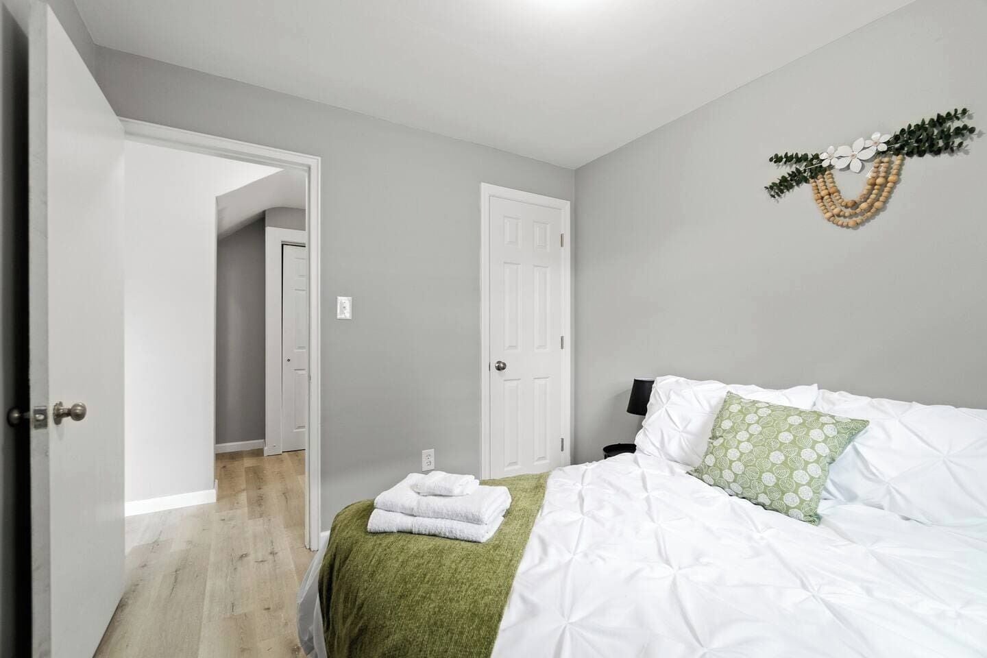 Un dormitorio con cama, una manta verde, almohadas blancas, un adorno en la pared y una puerta que da a otra habitación.