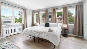 Et soveværelse med seng, radiator, et vindue med gardiner, et billede på væggen og udsigt til grønne områder.