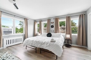 Et soveværelse med seng, radiator, et vindue med gardiner, et billede på væggen og udsigt til grønne områder.
