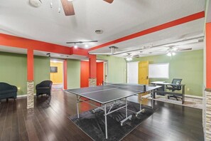 Una habitación con mesa de ping pong, dos sillas, un escritorio con computadora y un ventilador de techo.