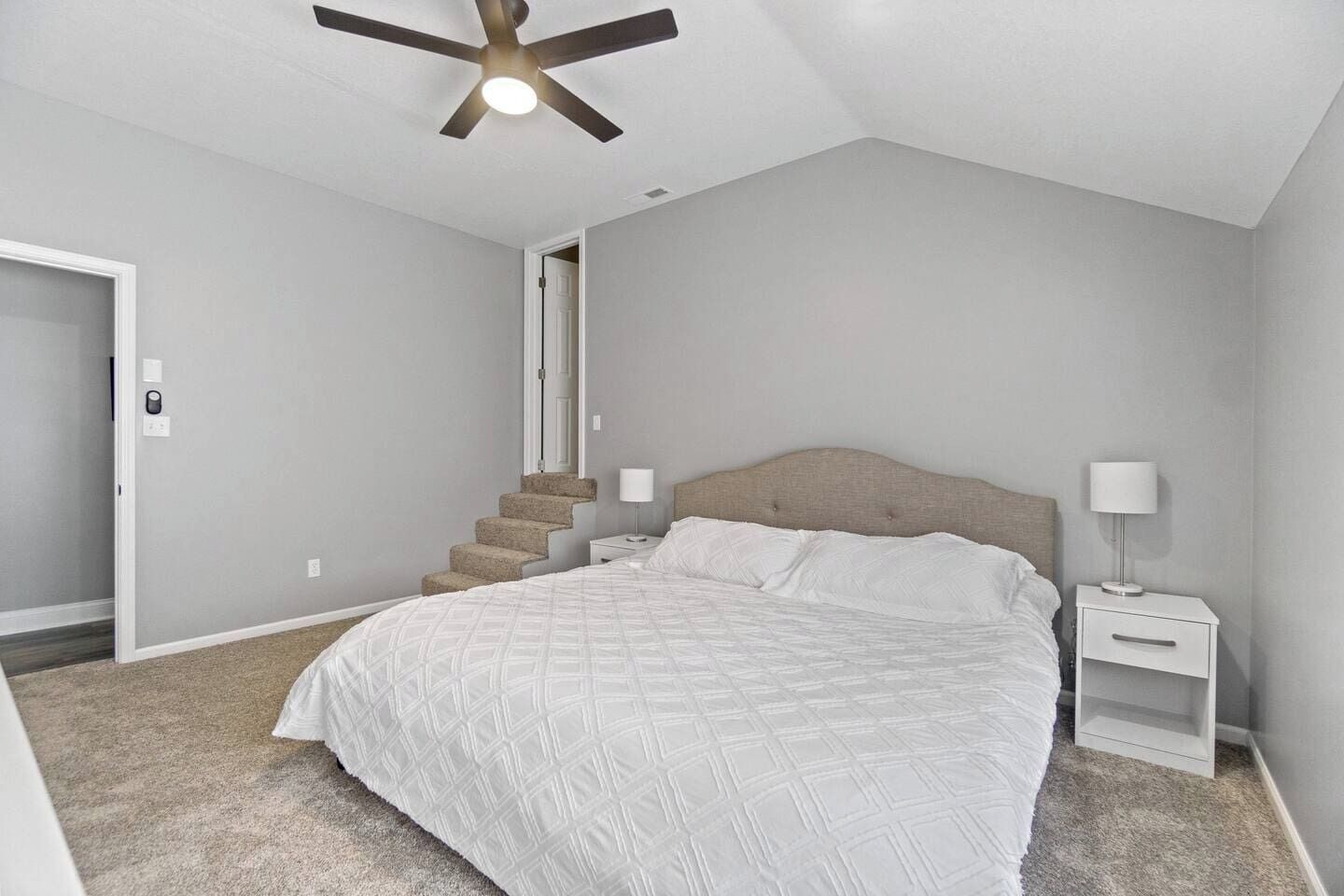 A bedroom with a bed, nightstands, a ceiling fan, a staircase, and a door.